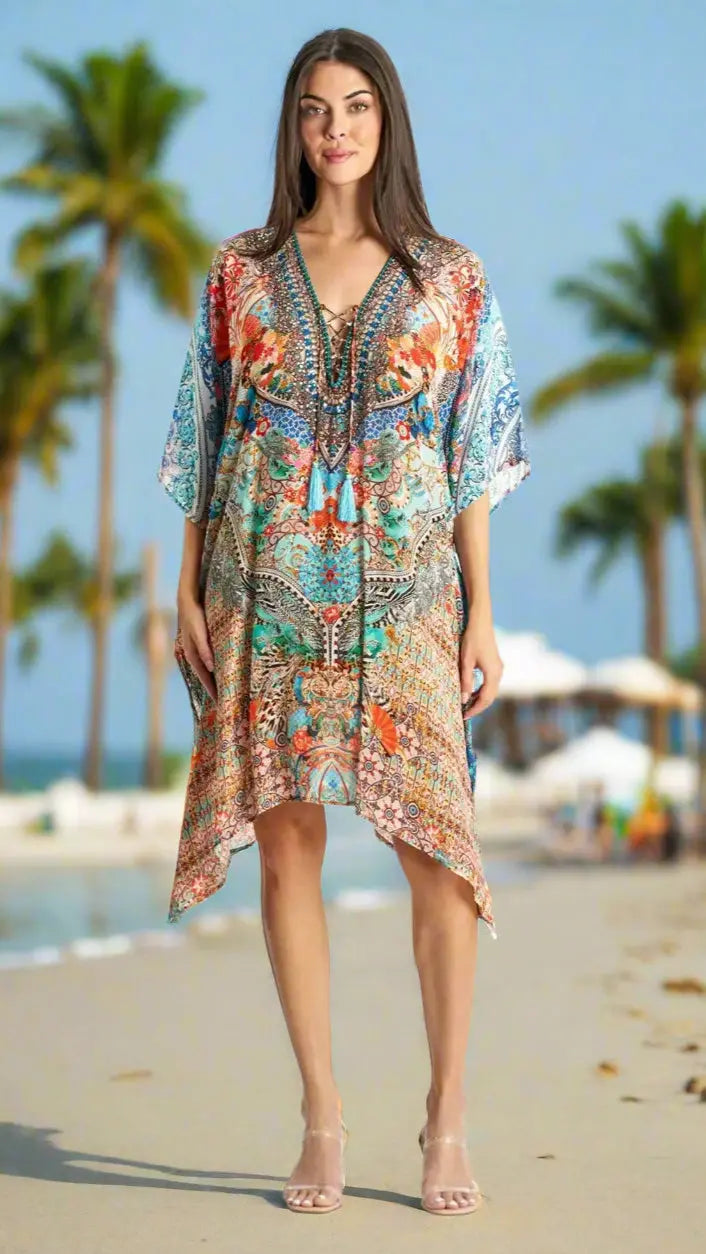 On a palm-lined sandy beach, a woman in a La Moda Clothing Caftans For Women wears a colorful, patterned, loose-fitting V-neck dress with a knee-length hem. She has long hair and open-toed heels; blurred beach chairs and umbrellas are behind her.