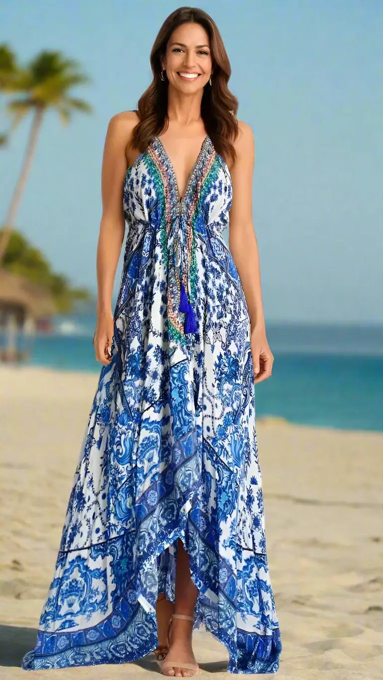 A woman stands barefoot on a sandy beach, smiling in the Casual Halter Maxi Dress by La Moda Clothing—a long blue-and-white patterned halter dress with colorful accents. The ocean, sky, palm trees, and a thatched hut blur in the background.