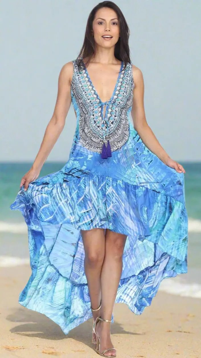 A woman smiles as she walks barefoot on the beach in a flowing Lamodaclothing Casual Summer Dress, featuring a sleeveless blue and white pattern and high-low hem. The ocean and sky blur in the background as she holds her dress's sides.