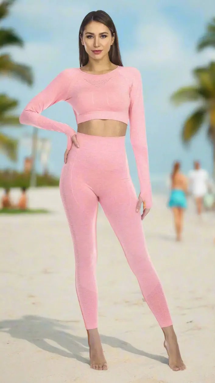 A woman stands barefoot on a sandy beach, wearing La Moda Clothings’ Club Moda Activewear Seamless Leggings & Joggers in light pink. Palm trees, blue sky, and blurred people in summer attire appear in the background as she faces forward and smiles slightly.