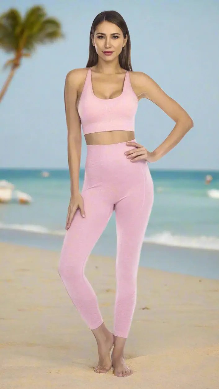A woman stands barefoot on a sandy beach, posing confidently in La Moda Clothings' Club Moda Rib Detail Seamless Gym Leggings and matching light pink sports bra, with the ocean, palm trees, and blue sky in the background.