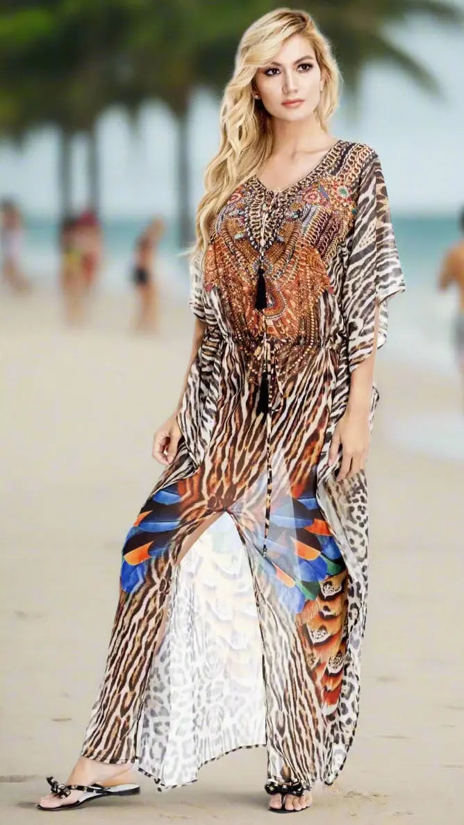 A woman with long blonde hair wears the Lamodaclothing Club Moda Women's Luxuriously Soft Maxi Kaftan, featuring bold animal-print feather patterns. She stands barefoot on a sandy beach with palms in the background, looking relaxed.