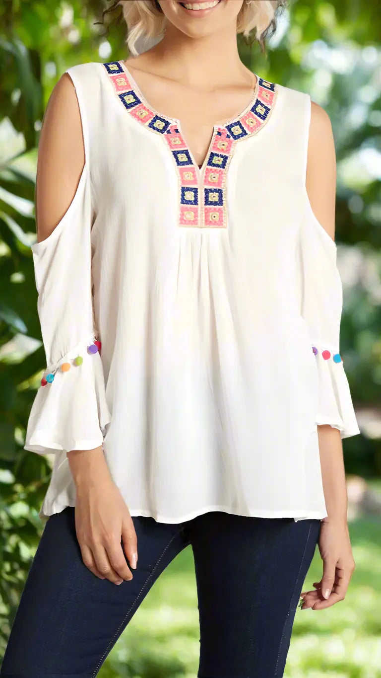 A woman wears the La Moda Clothing Cold Shoulder Beach Top—a loose white blouse with flared sleeves, colorful pom-poms, a square embroidered neckline, and open shoulders—paired with dark jeans. Blurred greenery is in the background.