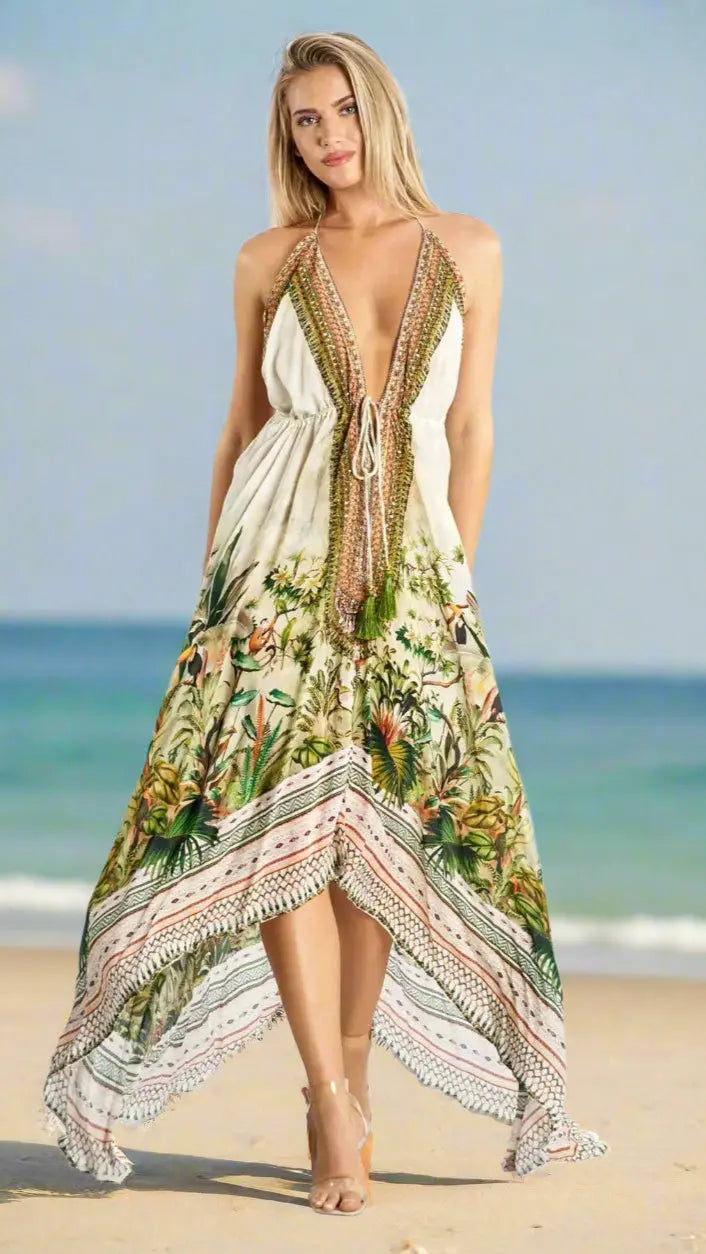 A woman stands on the beach in a white, sleeveless Convertible 3 Way Resort Maxi Dress by La Moda Clothings with a tropical floral pattern and beads. She wears beige sandals, her blond hair down, smiling softly with hands in her dress pockets.