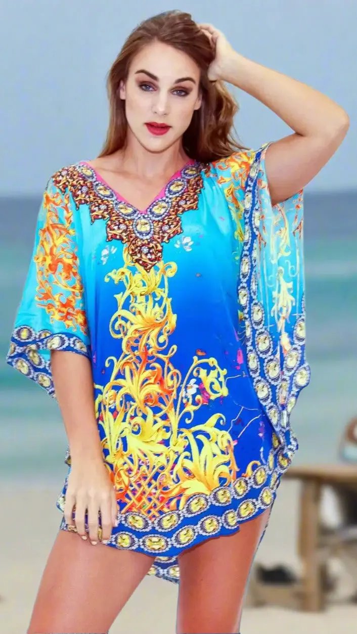 A woman with fair skin and long brown hair stands on a beach in the Designer Resort And BeachWear Kaftan by lamodaclothings—a vibrant blue and yellow tunic with intricate designs. She wears red lipstick, touching her hair, with the sea softly blurred behind her.