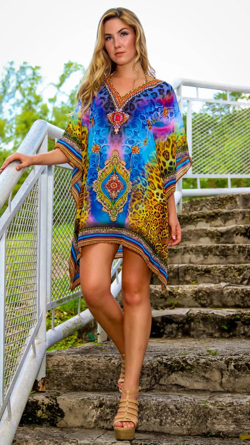 A woman with long blonde hair stands outdoors on stone steps, holding a white railing. She wears the Designer Resort Beachwear Kaftan by lamodaclothings—blue, yellow, and animal print with ornate designs—and beige wedge sandals. Green trees are behind her.