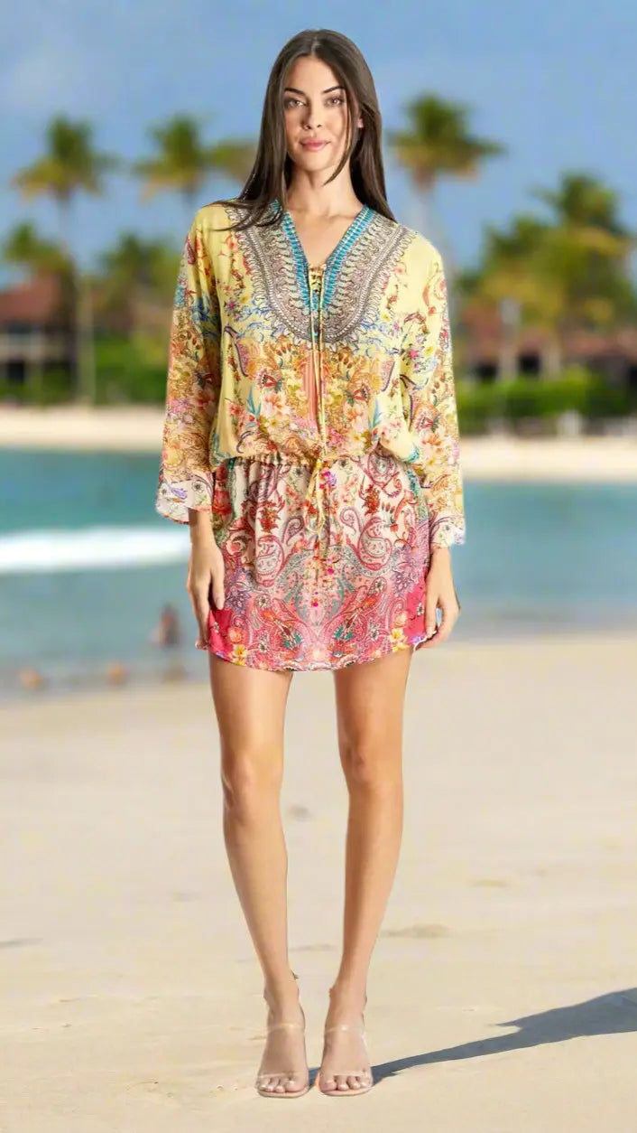 A woman stands on a sandy beach with palm trees and the ocean, smiling and facing forward. She wears the colorful Drawstring Tunic Dress by La Moda Clothing, featuring long sleeves and a vibrant print, paired with nude heeled sandals.