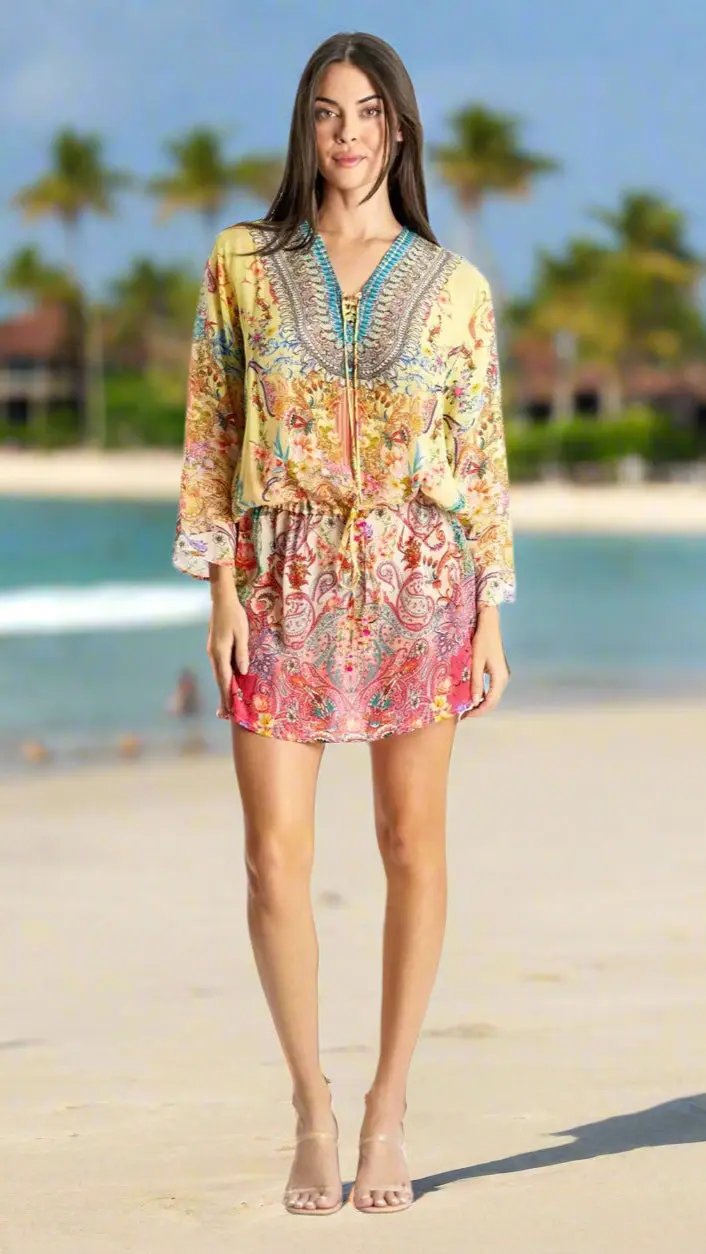A woman stands on a sandy beach with palm trees and the ocean, smiling and facing forward. She wears the colorful Drawstring Tunic Dress by La Moda Clothing, featuring long sleeves and a vibrant print, paired with nude heeled sandals.