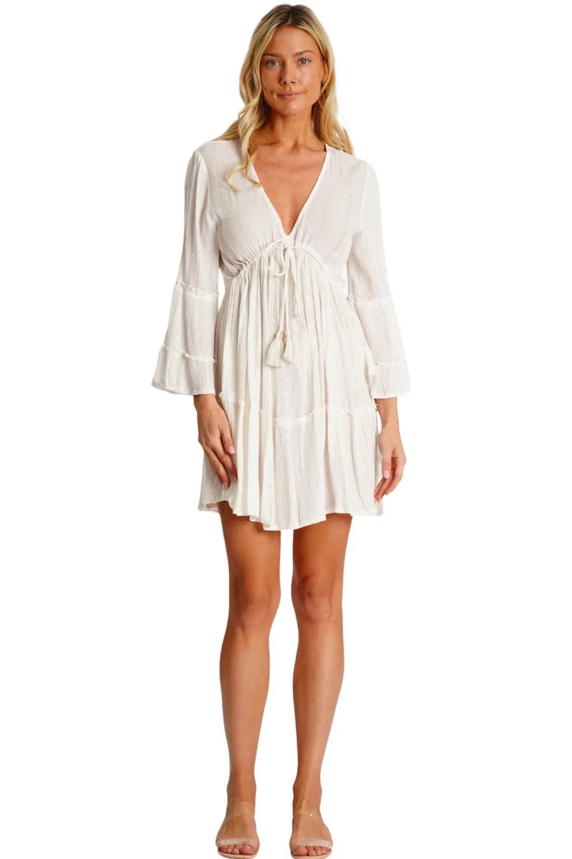 A woman wears the Dyed Mini Dress by La Moda Clothing—white, long-sleeve, v-neck with bust tie and ruffled hem—paired with clear heeled sandals, against a white background.