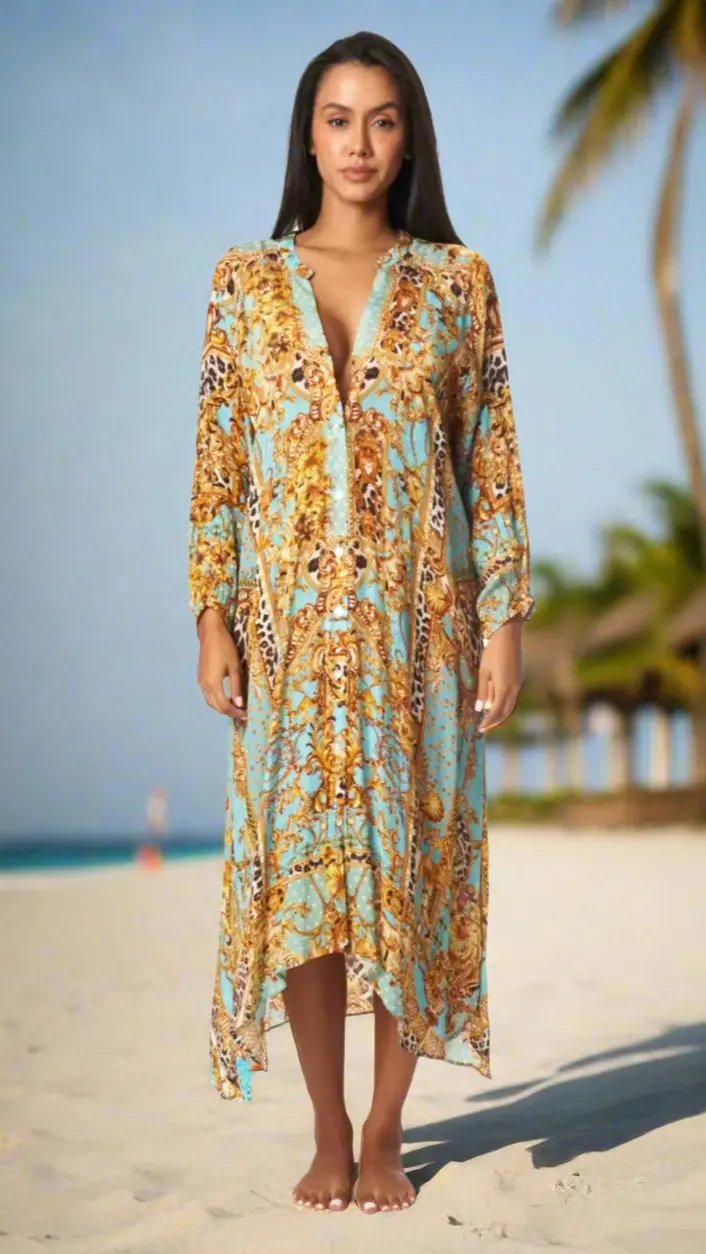 A woman stands barefoot on a sandy beach wearing the Easy Button Front Cover-Up Shirt Dress by La Moda Clothings, with its loose fit and vibrant blue and gold paisley pattern. Palm trees and a thatched hut blur in the sunlit background.