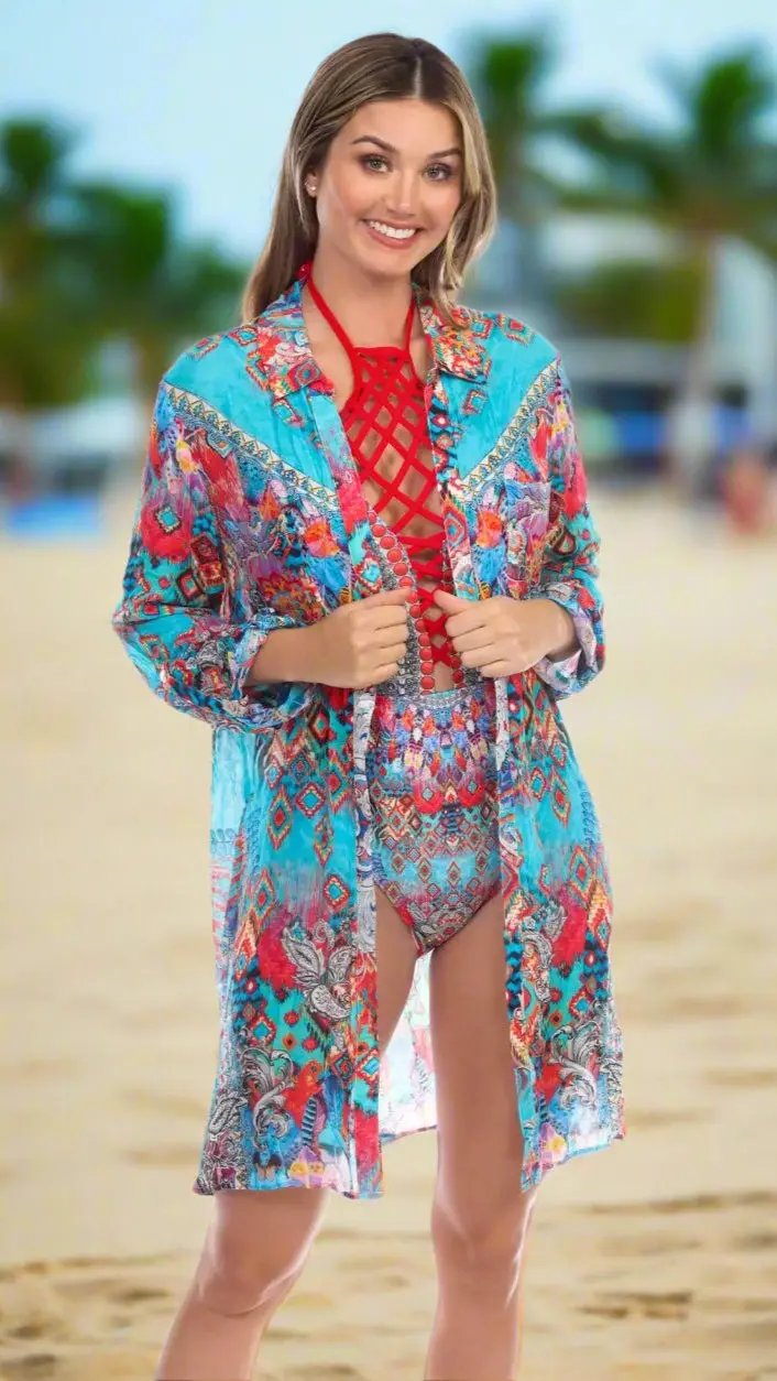 A smiling woman stands on a sandy beach, holding open the Easy Shirtdress Cover-Up by La Moda Clothings over her colorful swimsuit. Palm trees and a blue sky blur in the background as she enjoys the sun.