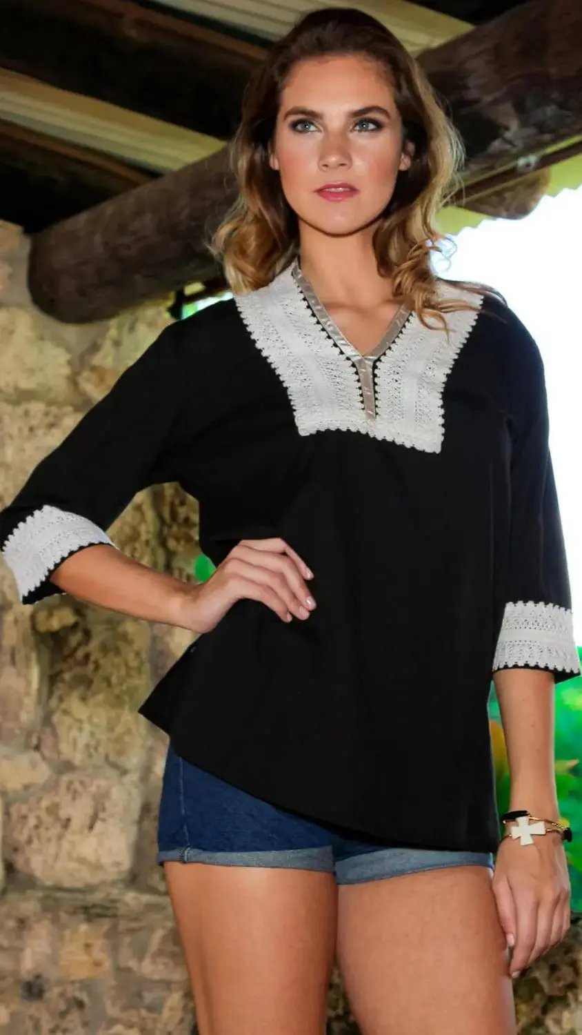 A woman with wavy brown hair poses indoors by a rustic stone wall, wearing the lamodaclothings Embroidered Long Sleeve Beach Tunic and Cover Up with white crochet details, paired with dark blue denim shorts.