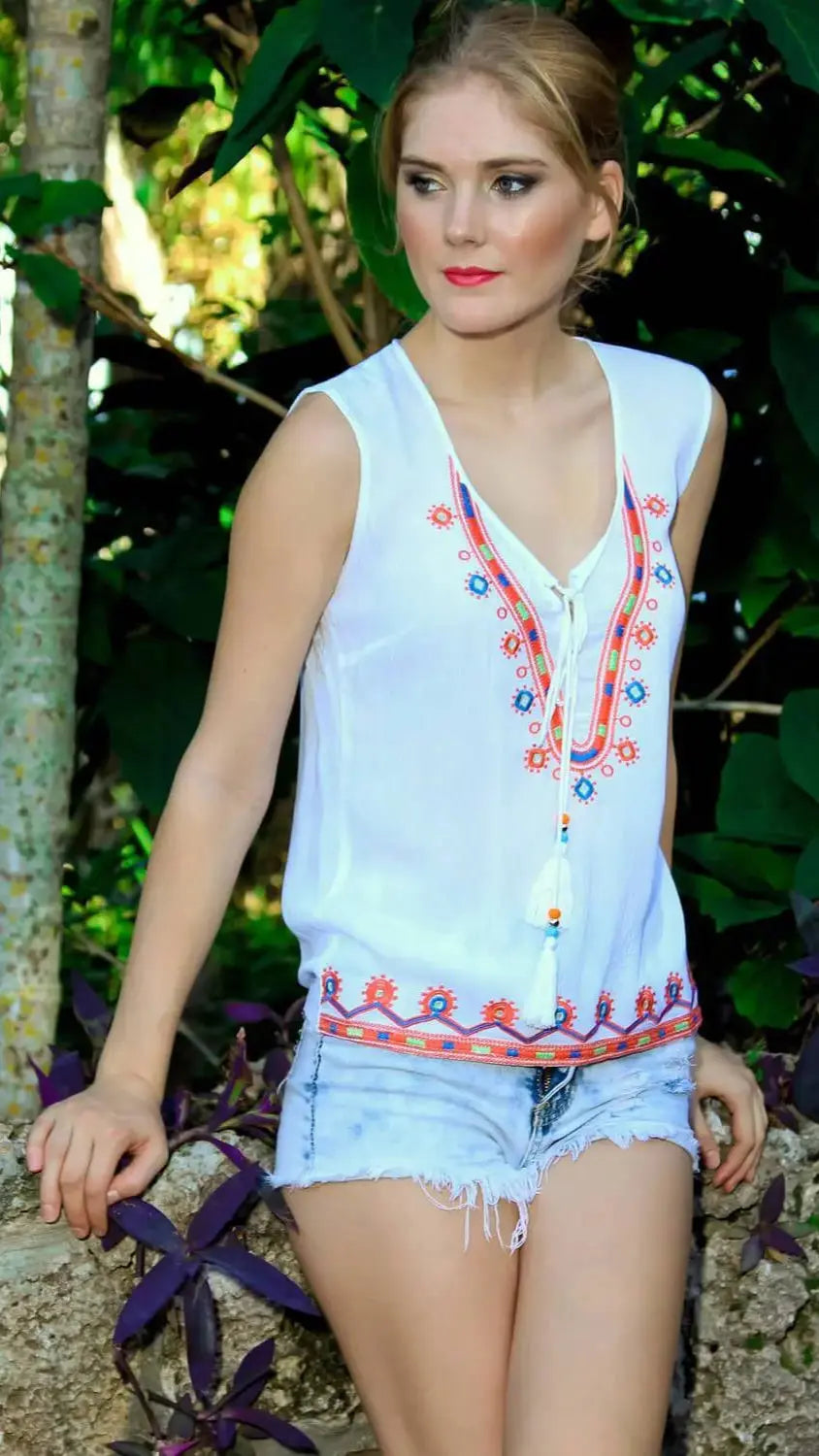 A young woman with light skin and blonde hair tied back sits outdoors on a stone ledge, surrounded by green leaves. She wears lamodaclothings’ Embroidered Top with Cross Lace and Tassels and light blue denim shorts, looking thoughtfully to the side.