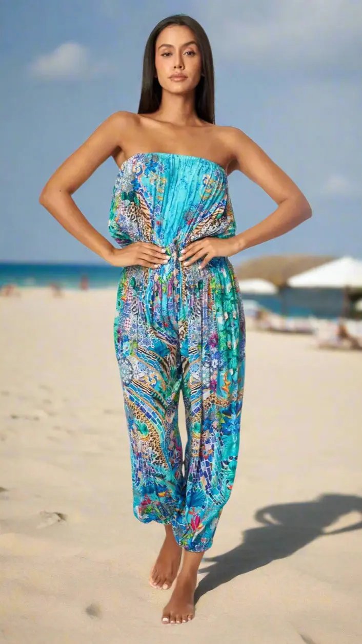 A woman stands barefoot on a sandy beach under a blue sky, wearing the lamodaclothings Garden Hippie Bohemian Jumpsuit with front pockets. She poses with hands on hips; her long hair flows as the ocean and beach umbrellas blur in the background.