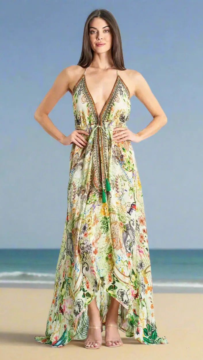 A woman stands on a sandy beach with the ocean behind her, wearing La Moda Clothing’s Halter High Low Maxi Resort Dress for Summer, featuring a plunging neckline, colorful florals, tie waist detail, and nude heels. Her arms rest on her hips.