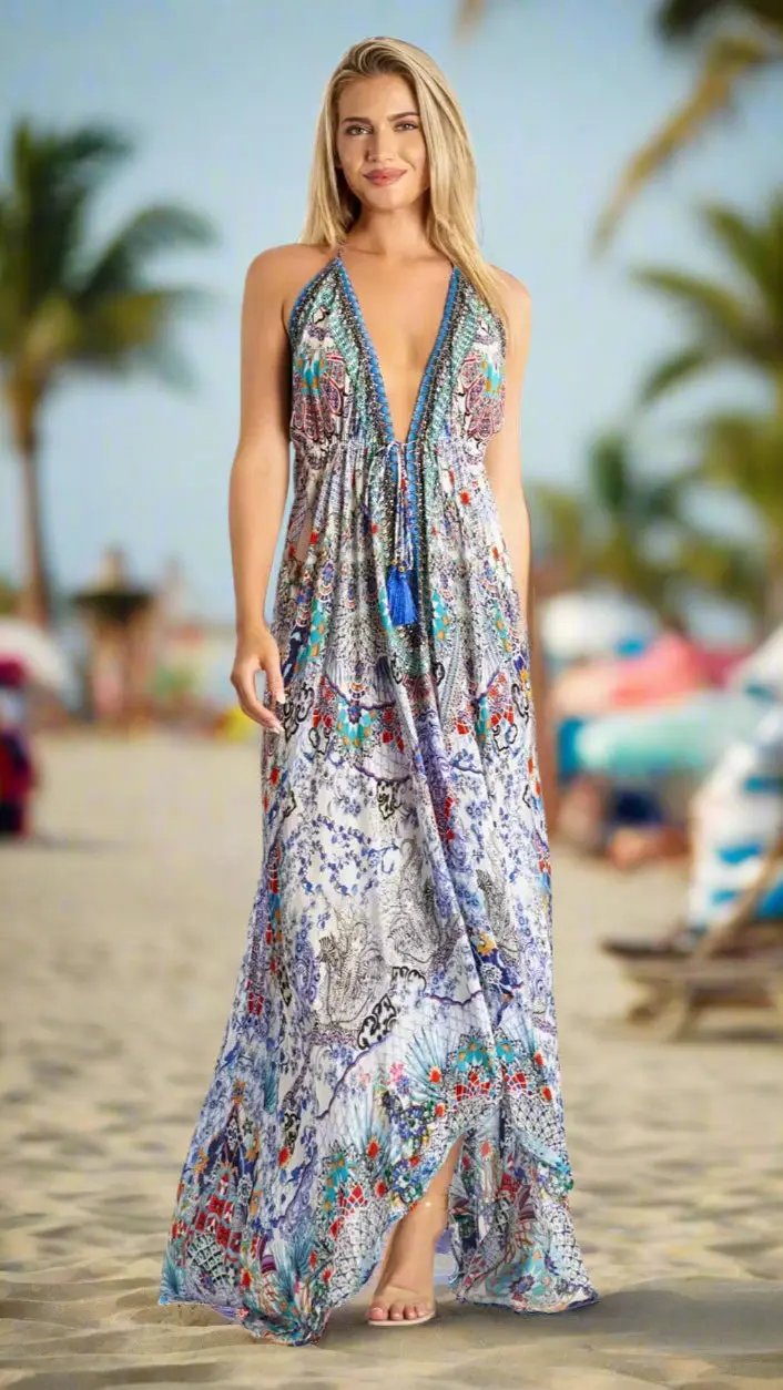 A woman stands barefoot on a sandy beach in a La Moda Clothing Halter Maxi Resort Dress, featuring a long, colorful design with a deep V-neck—perfect for tropical destinations. Palm trees and umbrellas complete the relaxed scene.