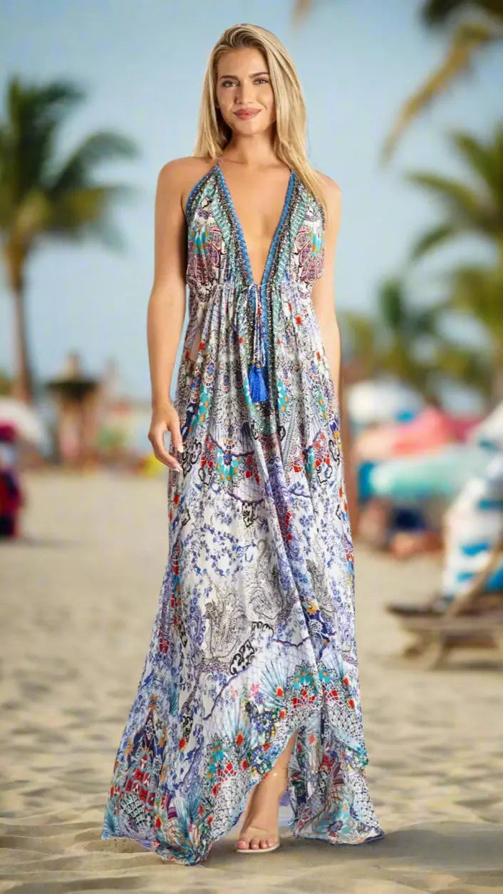 A woman stands barefoot on a sandy beach in a La Moda Clothing Halter Maxi Resort Dress, featuring a long, colorful design with a deep V-neck—perfect for tropical destinations. Palm trees and umbrellas complete the relaxed scene.