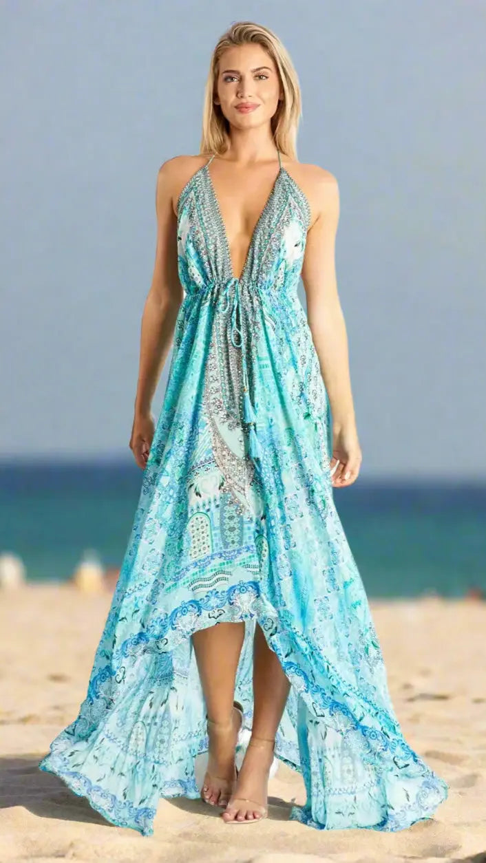 A woman stands on a sandy beach in the Halter Resort Maxi Dress by La Moda Clothing, featuring a light blue pattern, halter-neck, deep V neckline, and high-low hem. The blurred ocean and sky create a summery, relaxed vibe.