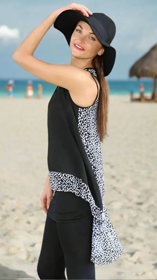 A woman with long brown hair smiles on a sandy beach, wearing Lamodaclothing's High Low Summer Tunics in animal prints and black leggings, holding a wide-brimmed black hat; people and a thatched hut appear near the sea in the background.