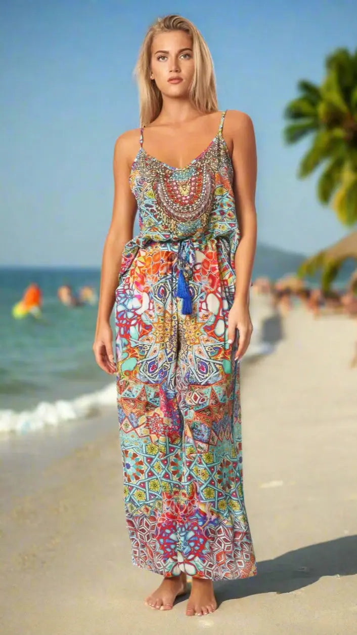 A woman with long blonde hair stands barefoot on a sandy beach wearing the Island Resort Co Ord Pant Set by La Moda Clothing. Palm trees and people swimming can be seen in the blurred background under a sunny sky.