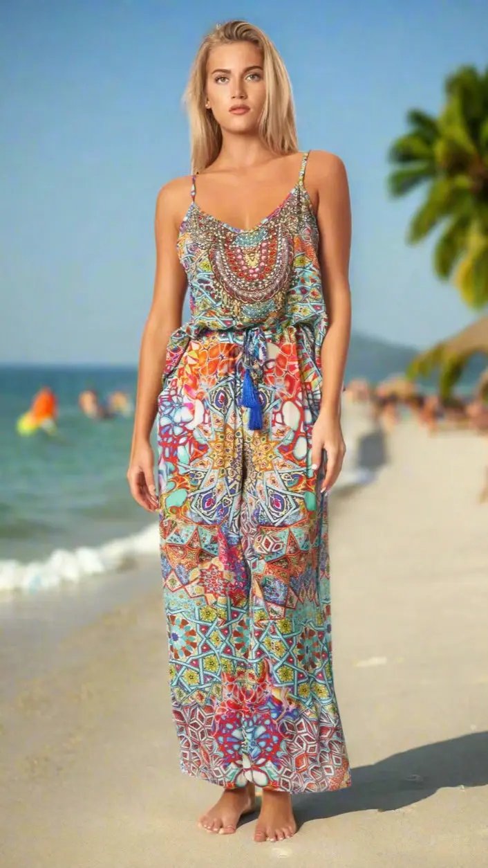 A woman with long blonde hair stands barefoot on a sandy beach wearing the Island Resort Co Ord Pant Set by La Moda Clothing. Palm trees and people swimming can be seen in the blurred background under a sunny sky.