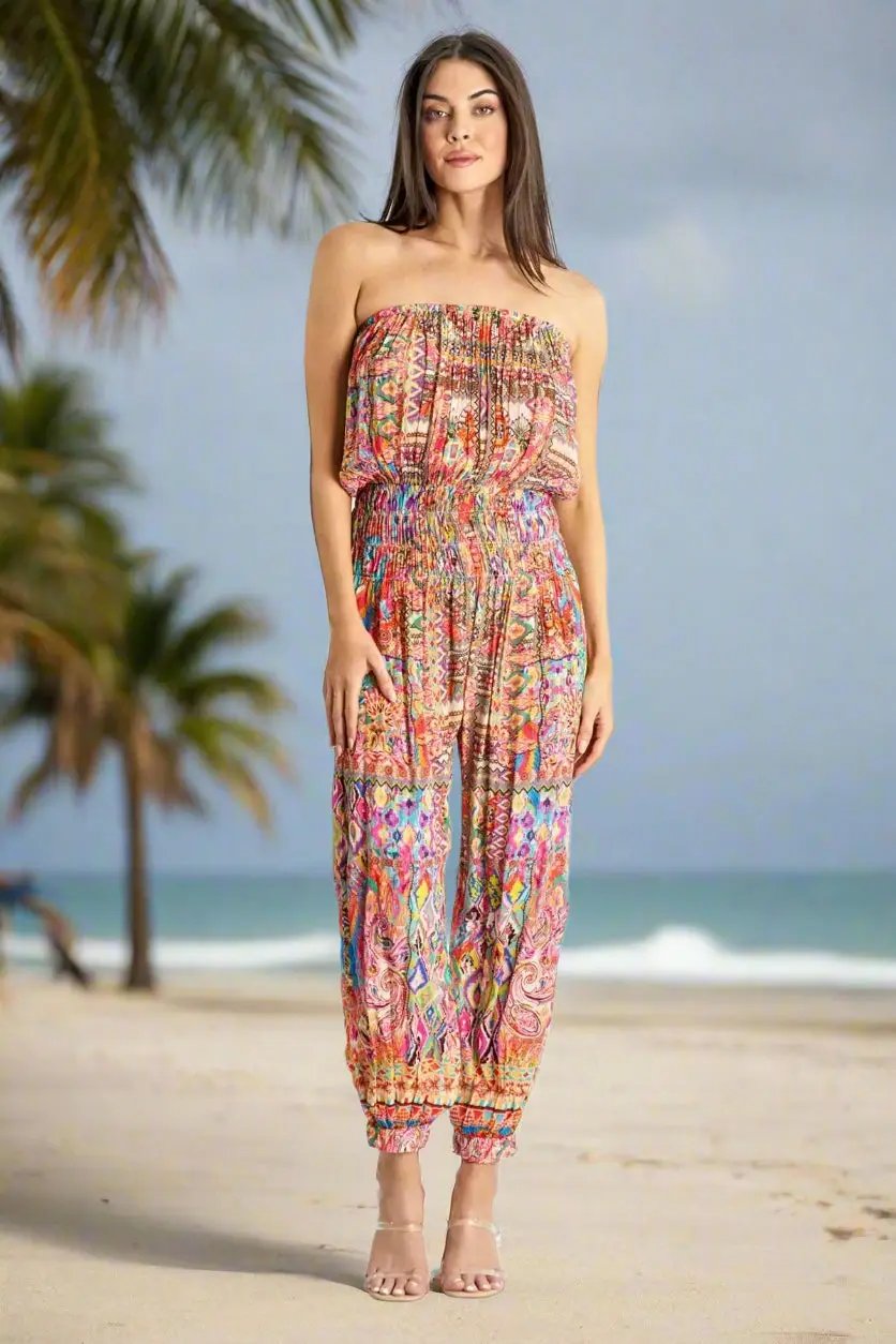 A woman with long brown hair stands barefoot on a sandy beach with palm trees and ocean waves, wearing a colorful La Moda Clothing Jumpsuit with a strapless, gathered top and ankles featuring a bright, intricate pattern.