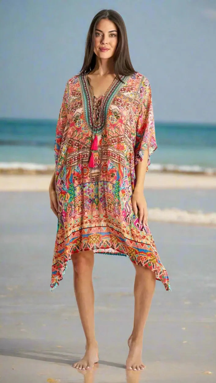 A woman stands barefoot on a sandy beach, wearing the La Moda Clothing Kaftan for Women with a colorful pattern, V-neck, and tassels. The clear sky and gentle ocean waves create a relaxed summery vibe in the background.