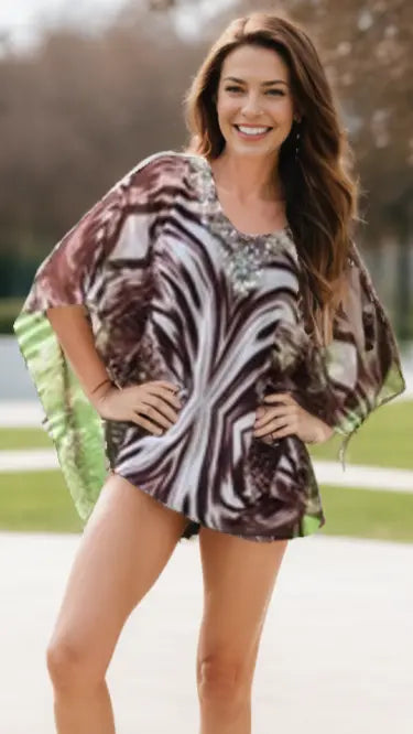 A woman with long brown hair smiles outdoors, wearing Lamodaclothing's "Beach to Bar" kaftan with a decorated neckline and abstract green, purple, and white swirls over shorts; blurred trees and a bright sky are behind her.