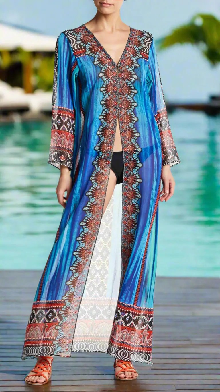 A woman by the pool wears the Kimono Cover Ups from La Moda Clothing—a luxury beach kimono with an open front, vivid blue and multicolored patterns, wide sleeves, and intricate red, white, and black designs over a black bikini. She sports orange sandals.