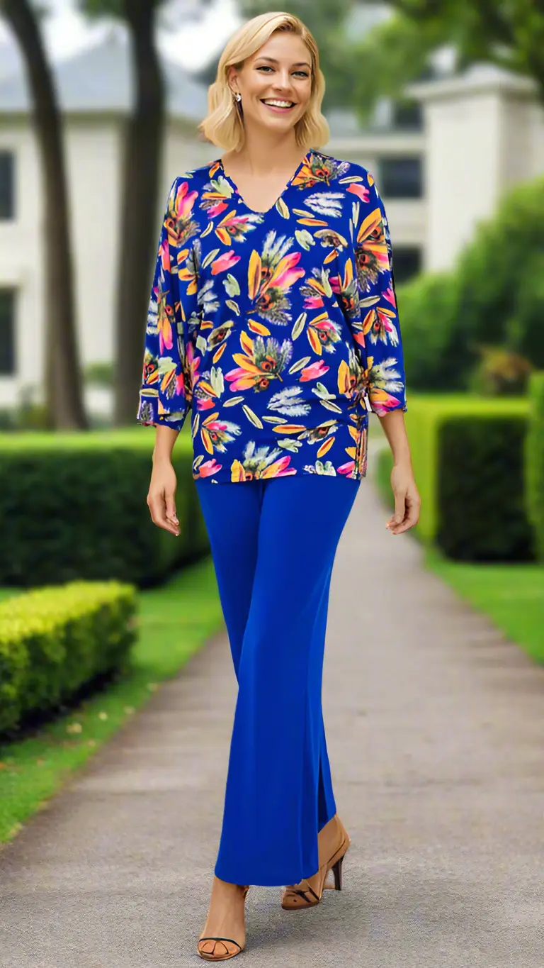 A smiling woman strolls a garden path in La Moda Clothing's Knit Bell Sleeve Resort Wear Tunic Top, featuring a blue floral print with orange, yellow, and pink flowers. Blurred trees and a white house appear in the background.
