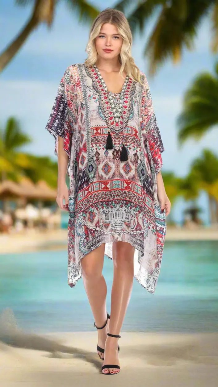 A blonde woman wears the La Moda Clothings Lightweight Rhinestone V Neck Printed Short Caftan with black sandals. She stands on a sunny sandy beach with palm trees and a blurred tropical resort in the background.