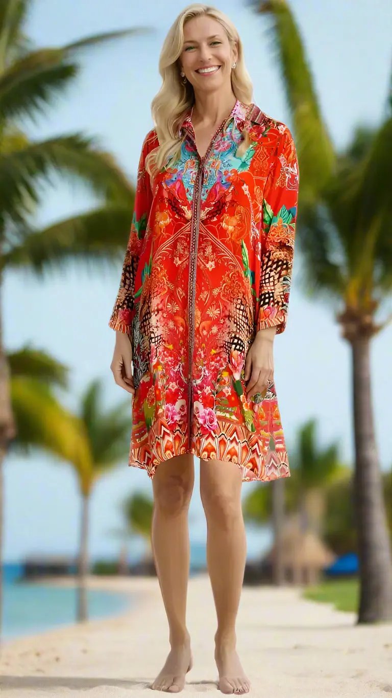 A smiling woman with long blonde hair stands barefoot on a sandy path lined with palm trees, wearing La Moda Clothing's Long Shirt Dress—a colorful, knee-length piece with vibrant red, pink, and orange floral patterns and long sleeves.