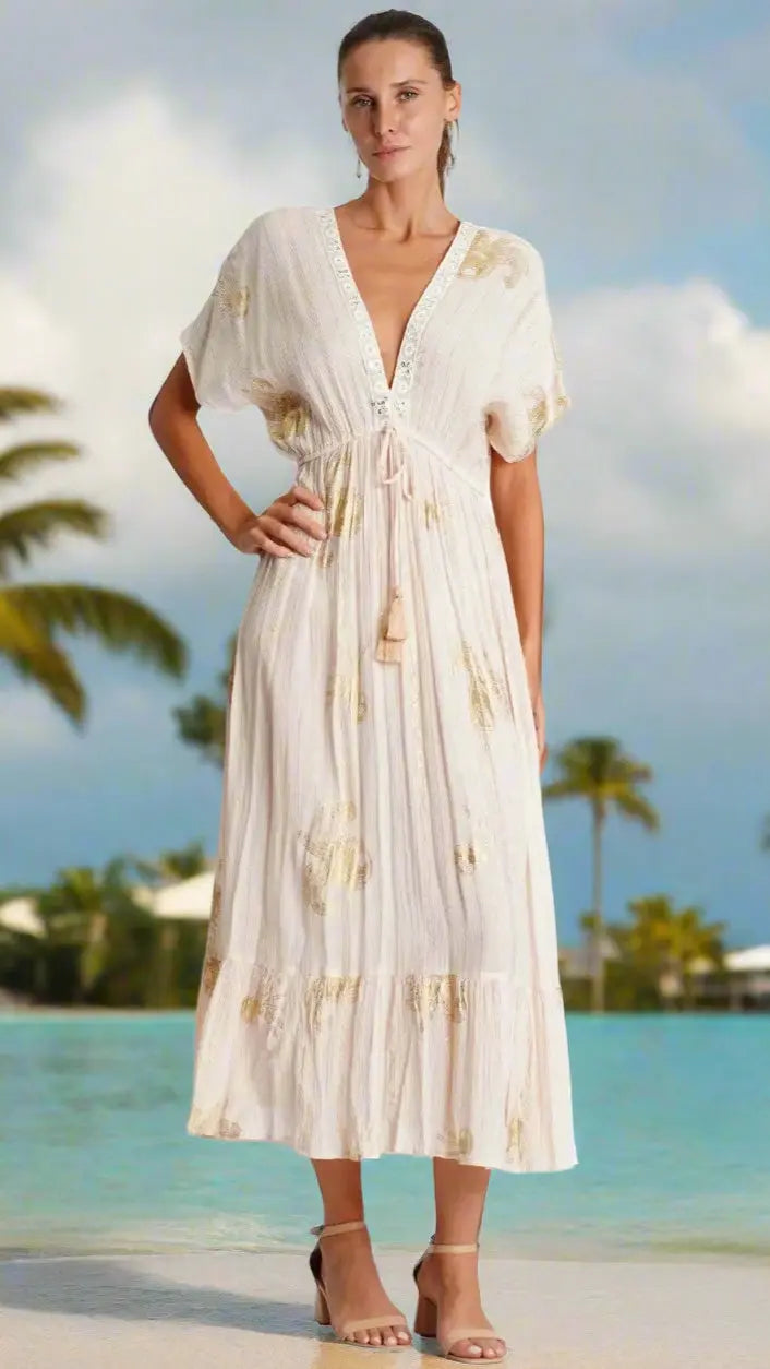 A woman stands by a turquoise pool wearing the Long White Maxi Dress by La Moda Clothing, featuring short sleeves, a V-neck with tassel ties, and flowing cream fabric. Her hair is pulled back, paired with nude heels under palm trees and blue sky.