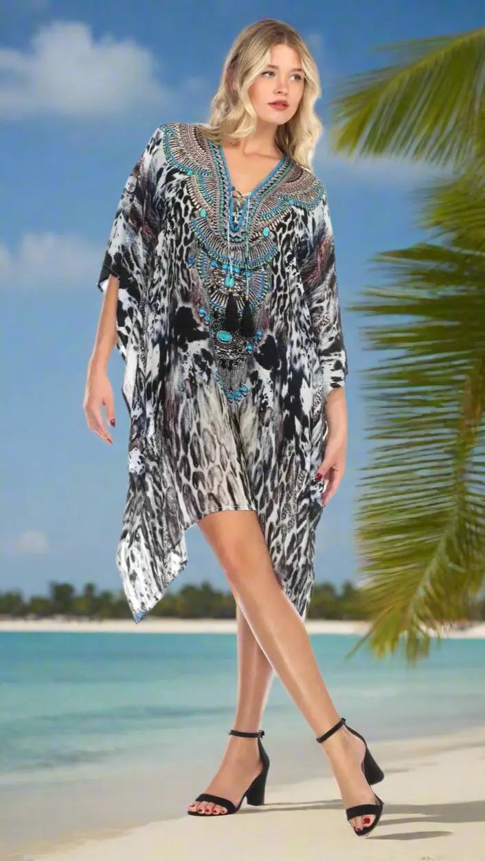 A woman with wavy blonde hair models the Luxury Embellished Caftans by lamodaclothings, featuring black, white, and gray animal print with turquoise beading. She stands barefoot on a tropical beach with palm fronds and a calm blue ocean behind her.