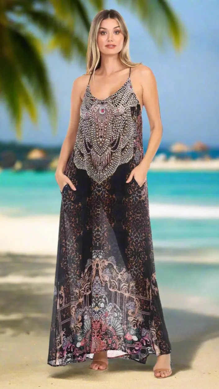 A woman stands on a tropical beach in the Maui Nights Elegant Maxi Pocket Dress by La Moda Clothings, a sleeveless, floor-length design in black, white, and pink viscose silk with ornate patterns and handy pockets. Blurred palms and sea form the background.