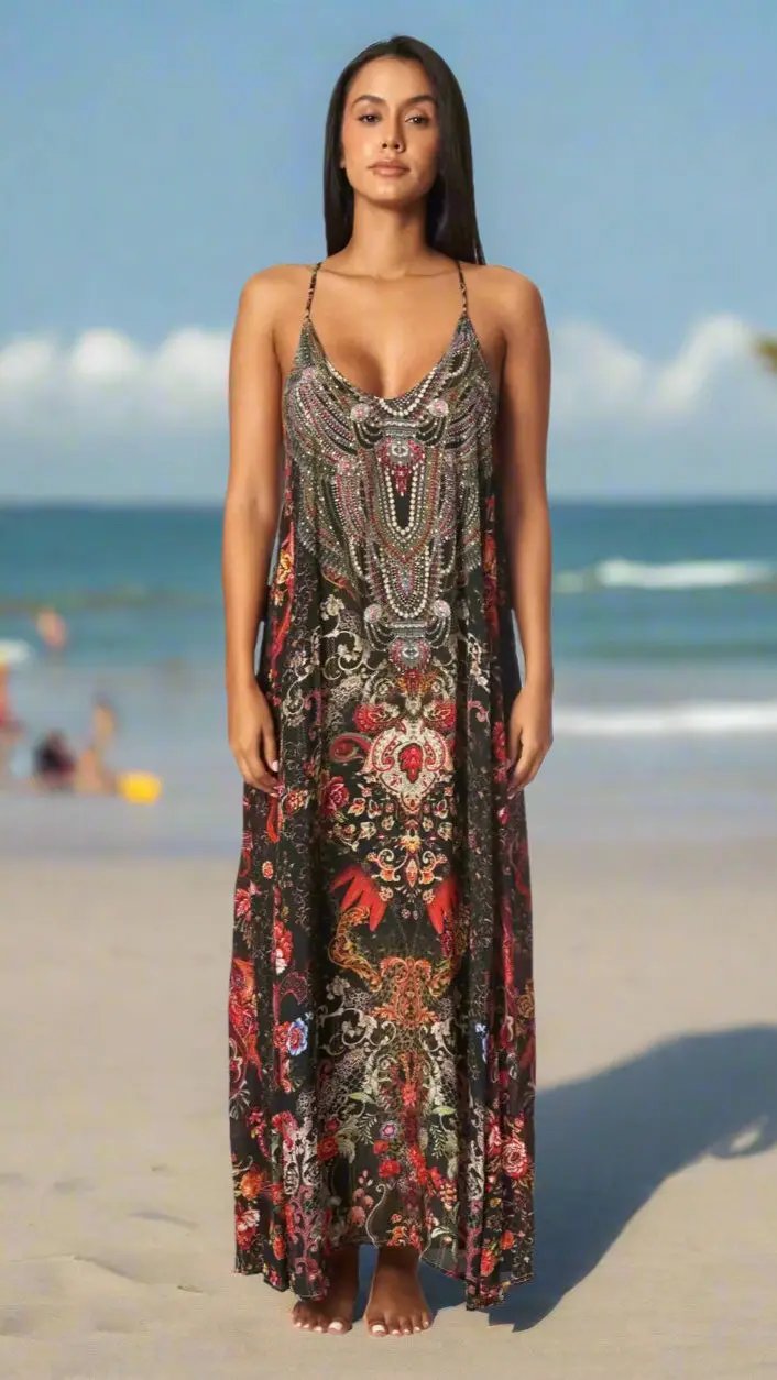A woman stands on a sandy beach wearing the Maxi Dress for Resort or Cruise by La Moda Clothings, featuring an intricate, colorful floral and paisley pattern. The calm sea and blue sky with clouds complete the serene scene.
