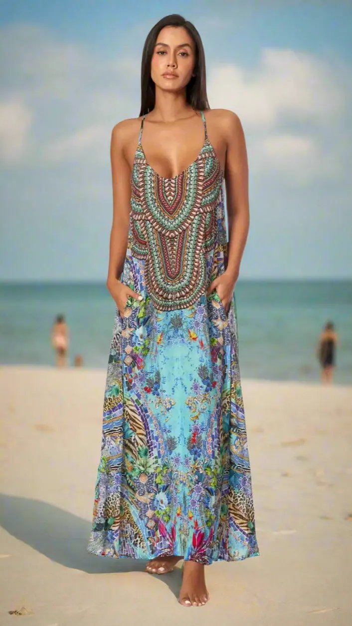 A woman barefoot on a sandy beach wears the La Moda Clothings Maxi Dress with Front Pockets for Resort or Cruise, featuring colorful red, blue, and green patterns. The sea and people appear in the background beneath a partly cloudy sky.