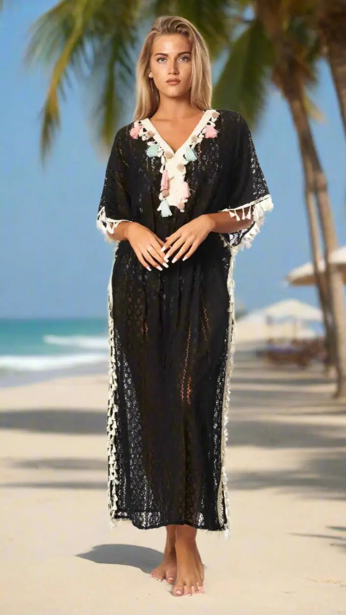 A woman stands barefoot on a sunny, tropical beach with palm trees and ocean behind her. She wears the Maxi Lace Kaftan Cover Up by La Moda Clothing—a black, semi-sheer dress with white trim, floral embroidery, and tassels.