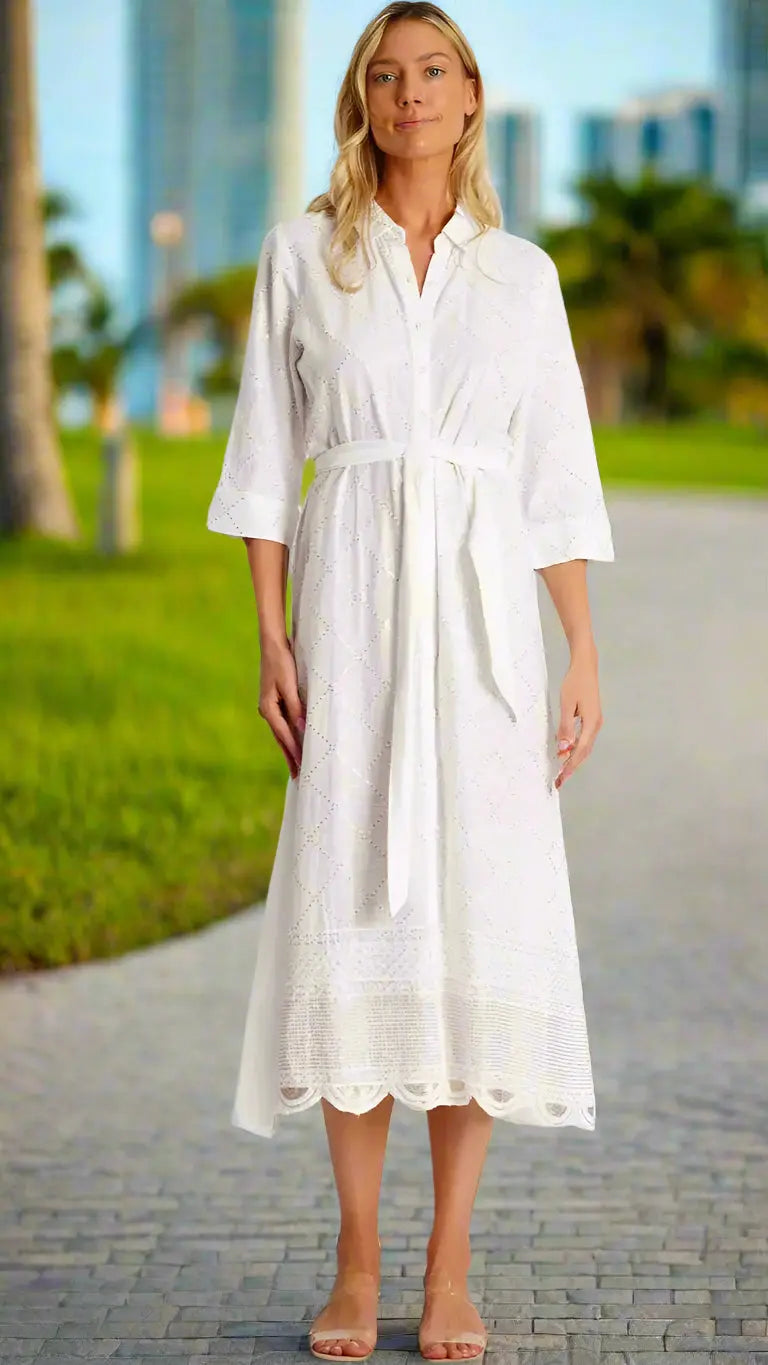 A woman stands on a sunny park path wearing La Moda Clothing's Maxi Shirt Dress—white, belted, with eyelet detail, three-quarter sleeves, and scalloped hem. She pairs it with sandals; greenery and city buildings blur in the background.