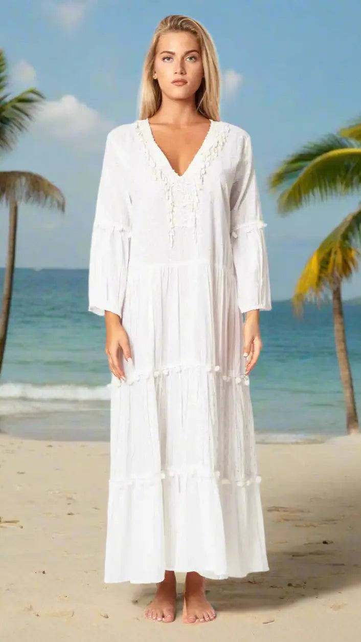 A woman with long blonde hair stands barefoot on a sandy beach in a La Moda Clothing Maxi Vacation & Summer Dress. The loose-fitting white dress features a V-neck and three-quarter sleeves, with palm trees and the ocean in the background.