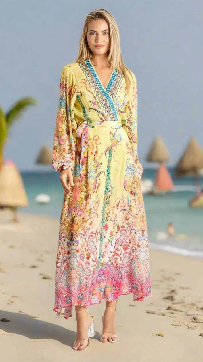 A woman stands barefoot on the sand in the Maxi Wrap Dress by La Moda Clothing—a long, flowing yellow dress with colorful florals and paisley, long sleeves, and a blue-trimmed V-neck. Calm sea, beach huts, and palms complete the scene.