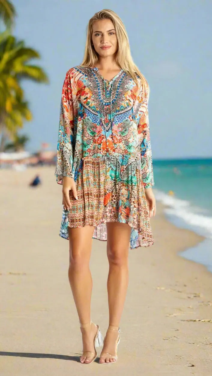 A smiling woman with long blonde hair stands on a sandy beach, wearing the La Moda Clothing Mini Dress for Beach Resort & Cruise Vacation. Palm trees and the ocean are in the background under a clear sky as she looks at the camera.