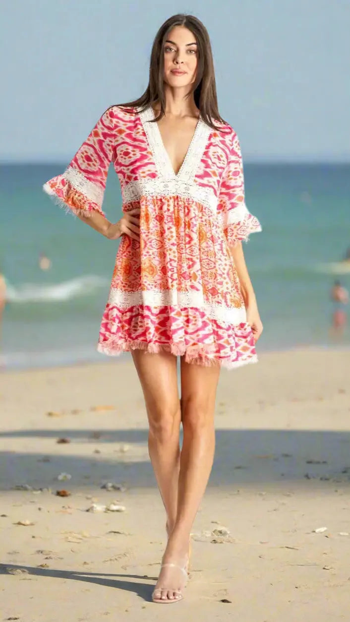 A woman stands on a sandy beach with the sea behind her, wearing the La Moda Clothing Mini Resort Wear Dress for Women—a short, loose fit featuring pink and orange tribal patterns, white lace trim, flared sleeves, and clear sandals.