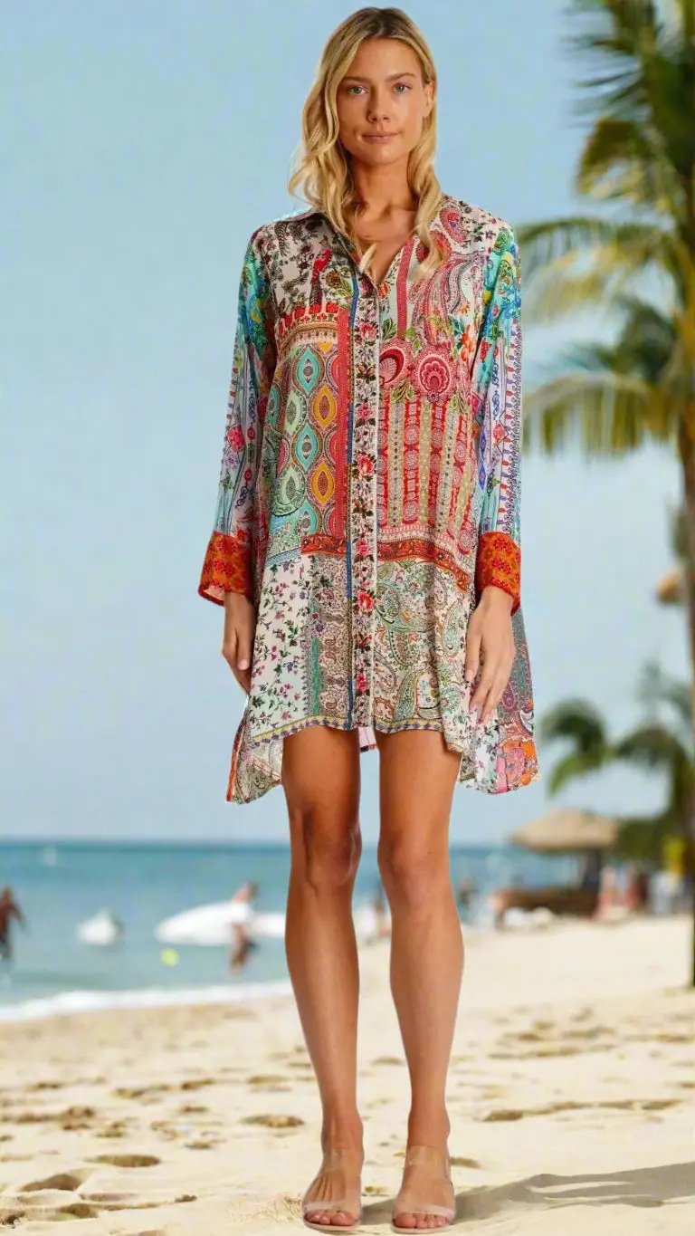 A woman stands barefoot on a sandy beach, wearing the La Moda Clothing MixMatch Print Long Tunic, featuring vibrant mixed patterns and red cuffs. Palm trees, sunbathers, and a blue sky with light clouds are seen in the background.