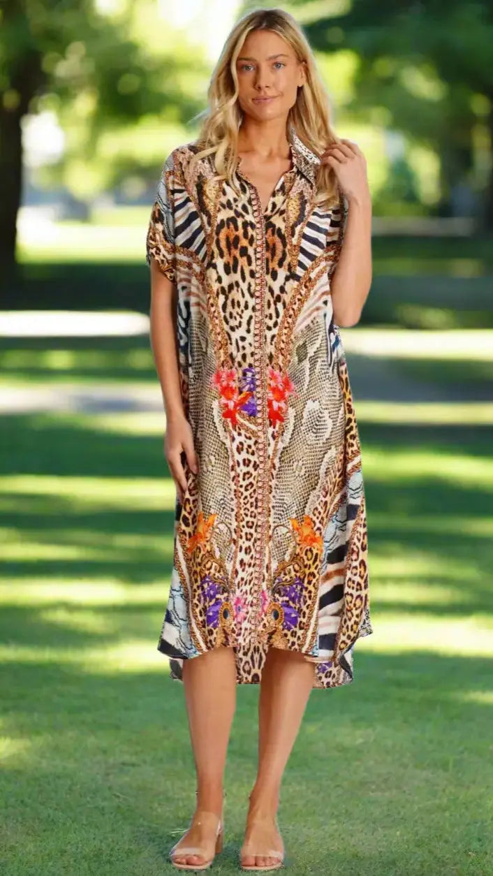 A woman with long blonde hair stands on grass in a park wearing La Moda Clothing's Mixed Animal Pattern Shirt Dress, featuring bold leopard, cheetah, and zebra prints with orange, red, and purple accents. She pairs it with neutral sandals.