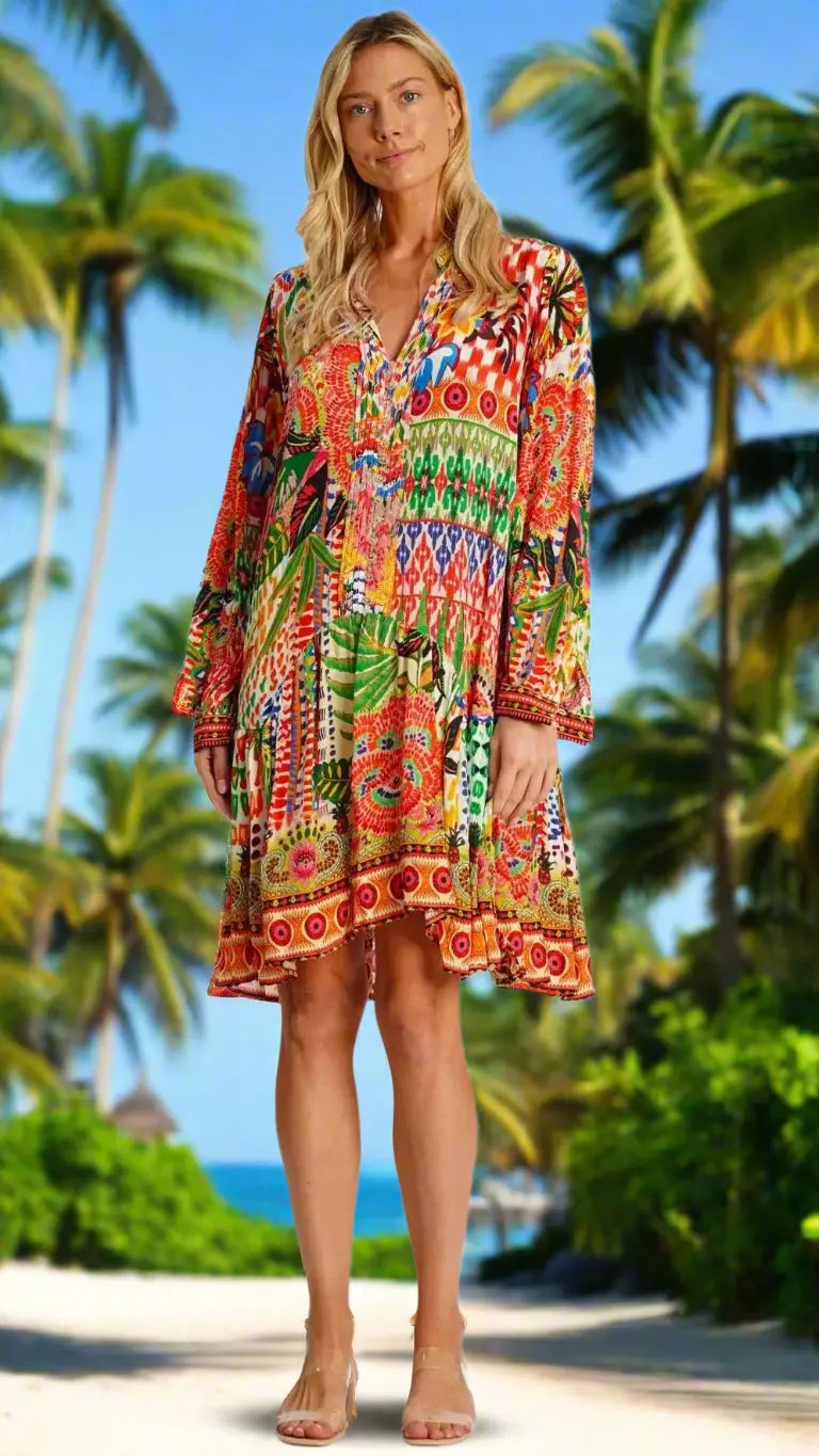 A woman with long blonde hair stands on a sandy beach with palm trees and the ocean, wearing the Natalie Tropical Shirtdress by La Moda Clothing—a colorful, patterned knee-length dress for a relaxed, summery look.