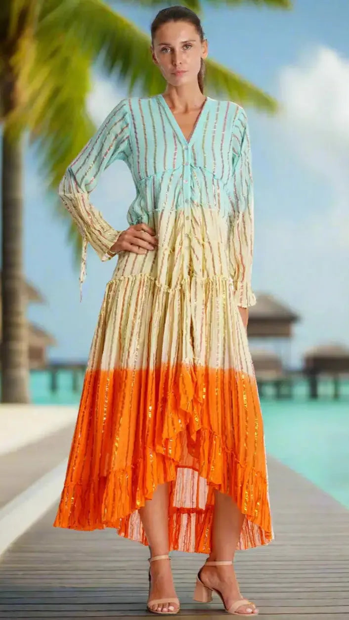 A woman stands on a boardwalk near water and overwater bungalows, wearing the Ombrey Tiered High Low Dress by La Moda Clothing, featuring blue, cream, and orange stripes. She has light brown hair pulled back and is wearing beige sandals.