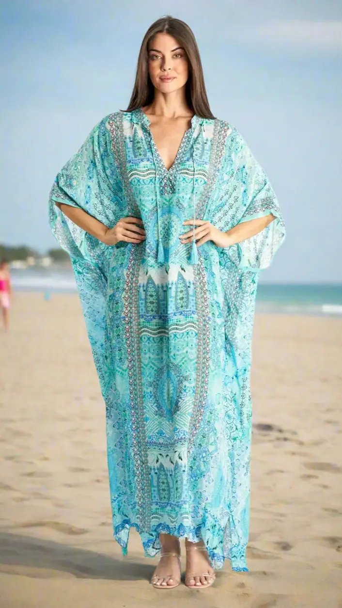 A woman with long brown hair stands barefoot on a sandy beach, wearing the Oversized Boho Maxi Caftan by La Moda Clothings. The flowing caftan features turquoise patterns and intricate designs, with the ocean and light blue sky in the background.