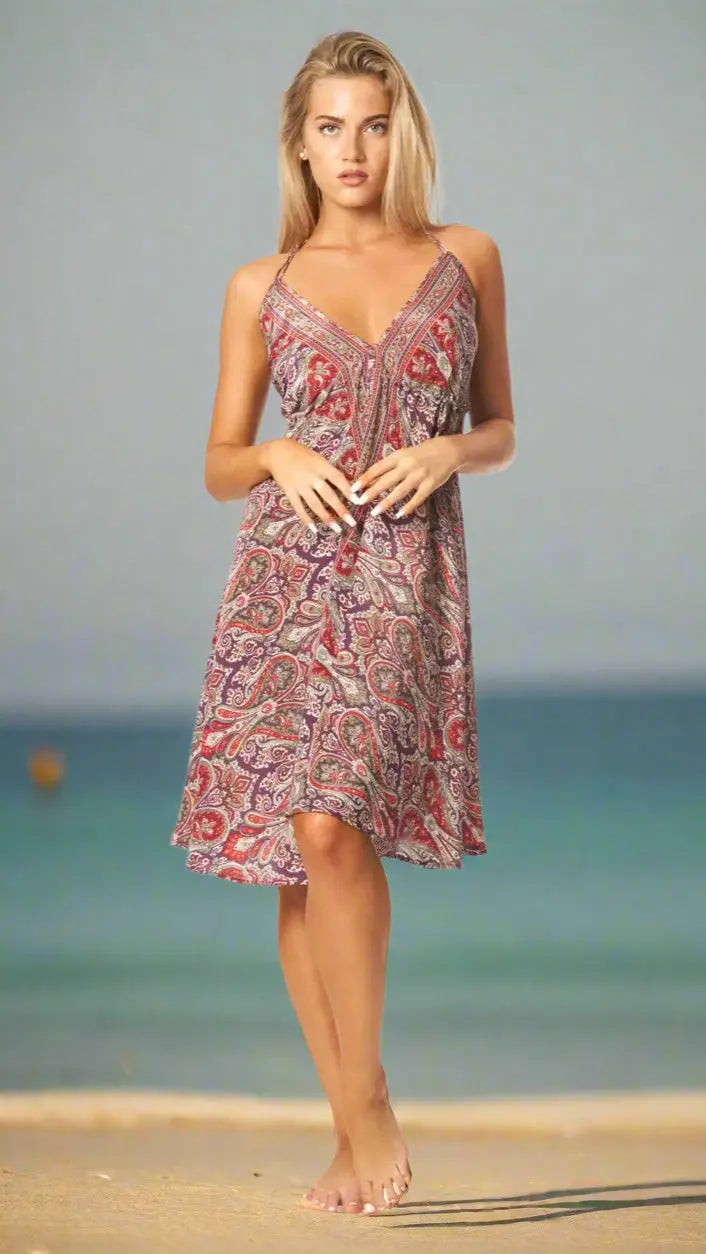 A woman stands barefoot on a sandy beach, wearing La Moda Clothing’s Positano Mini Boho Dress—sleeveless and loose-fitting with a colorful paisley pattern—perfect for Mediterranean cruises in a relaxed, summery setting.