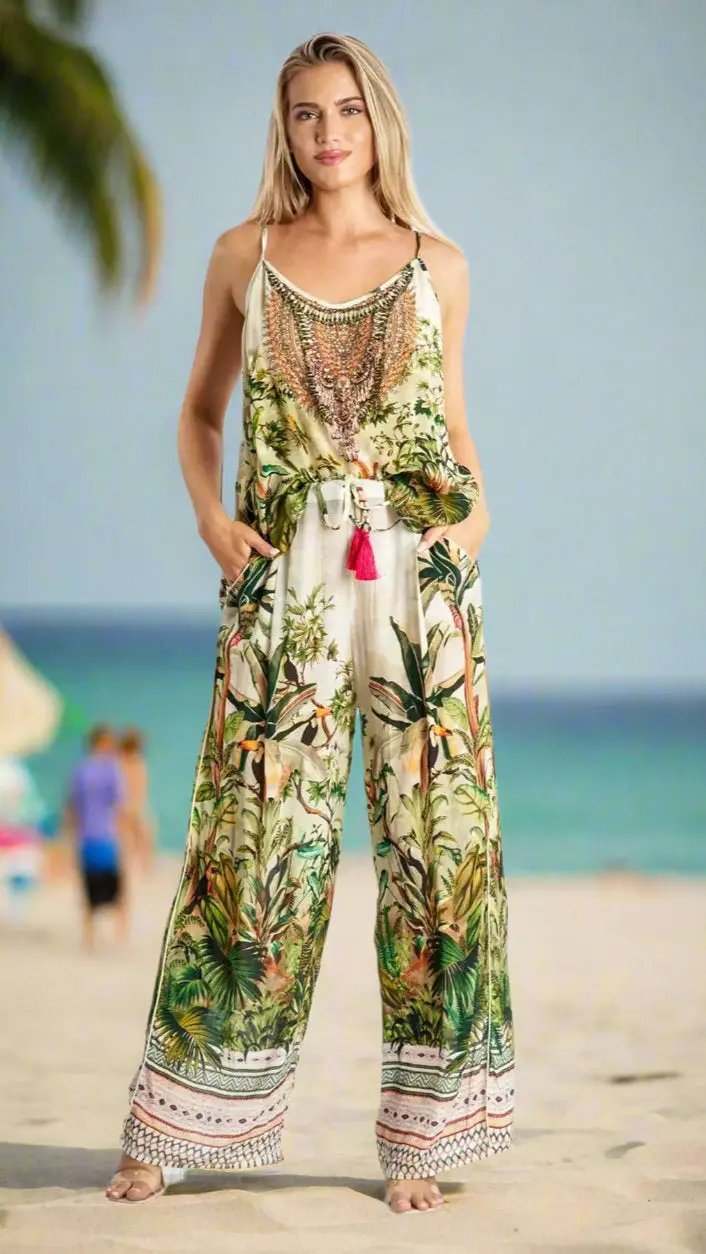 A woman stands barefoot on a sandy beach wearing the Resort Lifestyle Co Ord Pant Set by La Moda Clothing, a loose, sleeveless outfit with tropical botanical print and tassel accents. She gazes ahead with her hands in her pockets; the ocean is behind her.